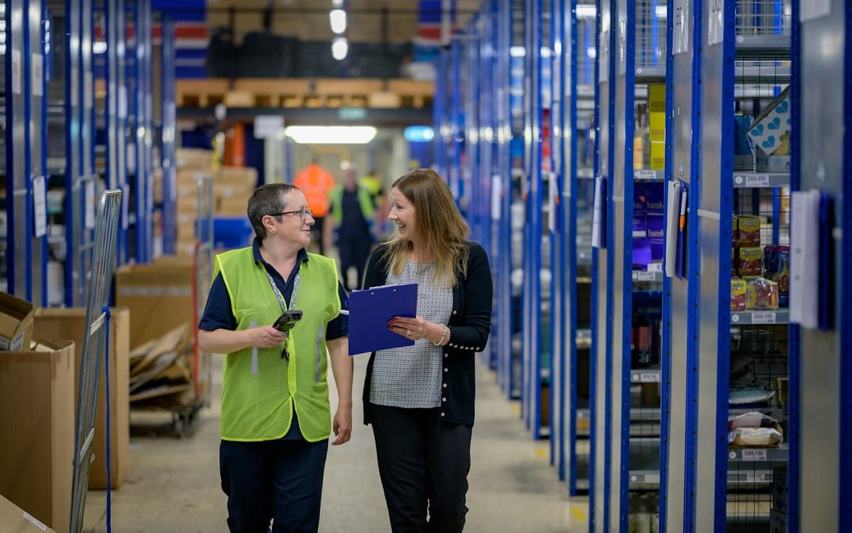 Two warehouse colleagues in hi-vis talking as they walk down a bright aisle of racked, labelled storage